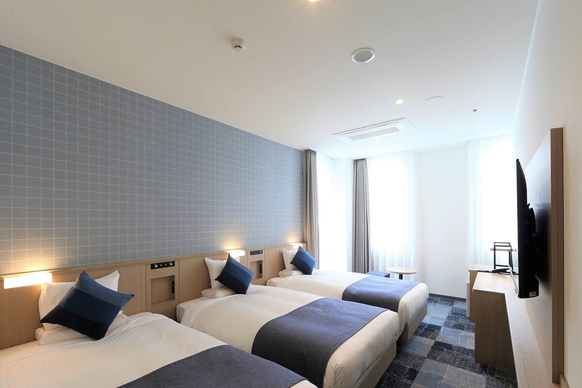 A hotel room with three beds, a desk, a flat-screen TV, and a large window with curtains.