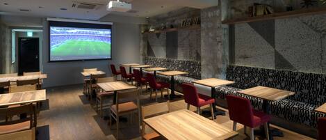 A modern restaurant with wooden tables and red chairs, a large screen displaying a sports event, and a stone wall with shelves.