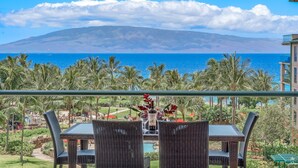 Un balcon avec des fauteuils en osier et une table, donnant sur une plage bordée de palmiers, avec une vue sur l’océan et une montagne.