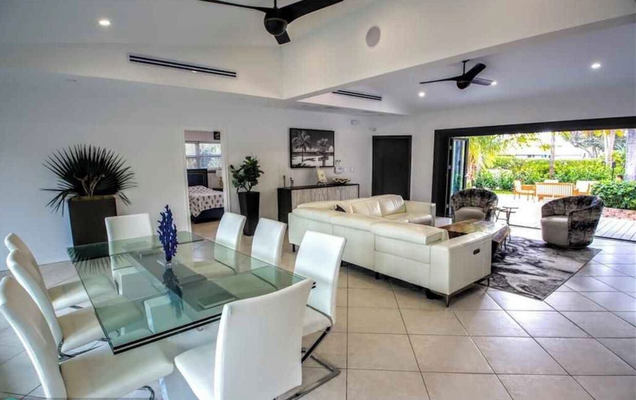 A modern living room with a glass dining table, white sofas, and a large window offering a view of the outdoors.