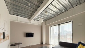 Una sala moderna con un televisor de pantalla plana montado en la pared, una mesa de centro de vidrio, un sofá oscuro con un cojín amarillo y ventanas grandes con vistas a la ciudad.