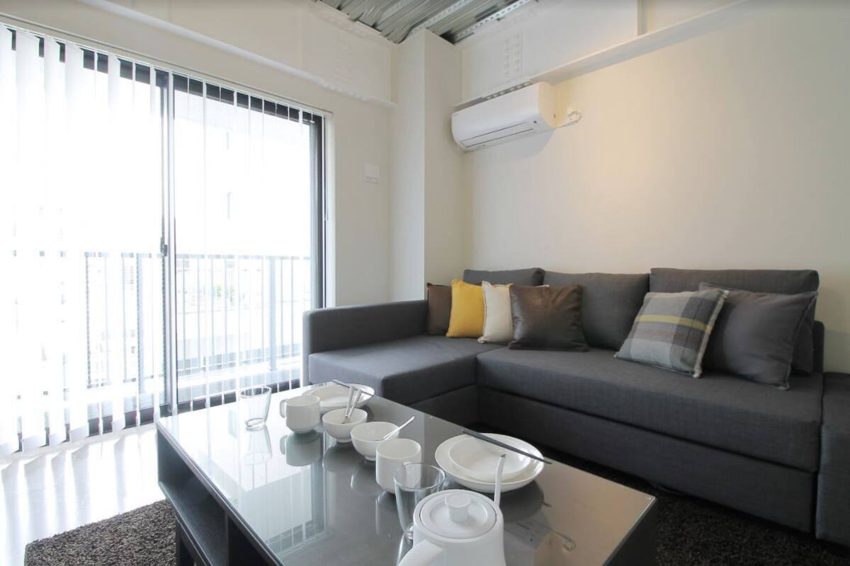 A modern living room with a grey sofa, a glass coffee table, and a sliding glass door leading to a balcony.