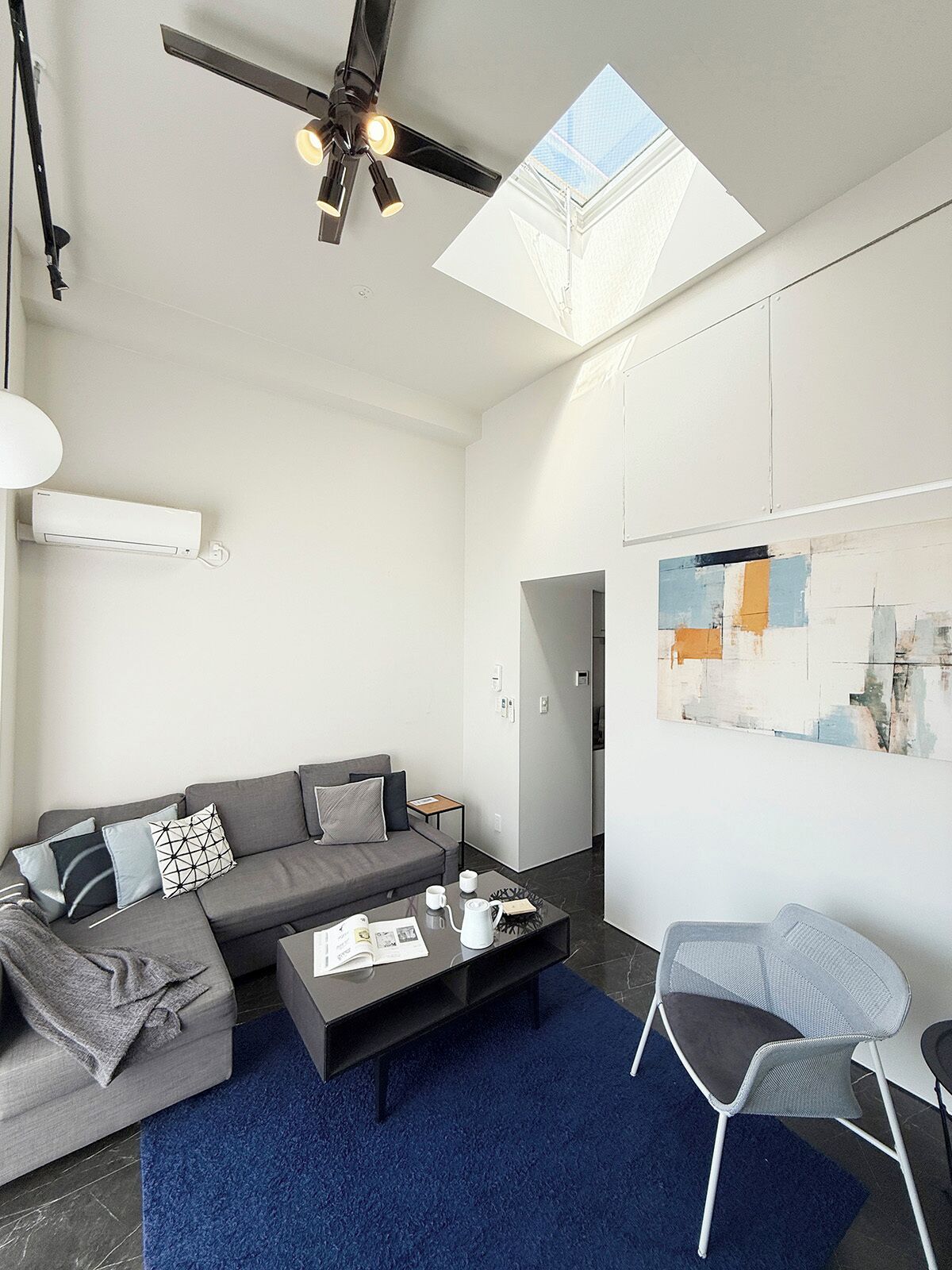 Ein modernes Wohnzimmer mit einem grauen Sofa, einem Couchtisch und einem blauen Teppich. Es gibt einen Deckenventilator, ein Oberlicht und eine wandmontierte Klimaanlage.