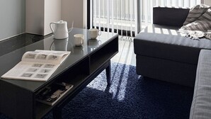 Una habitación de hotel moderna con una mesa de centro de vidrio, un sofá, un ventanal con persianas verticales y un techo con vigas a la vista.