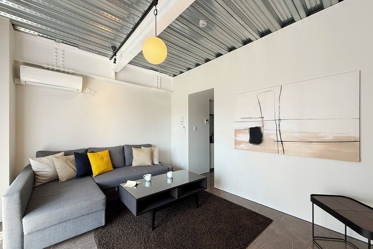 A modern living room with a grey sectional sofa, a coffee table, and a black rug. There is a painting on the wall and a pendant light above the sofa.