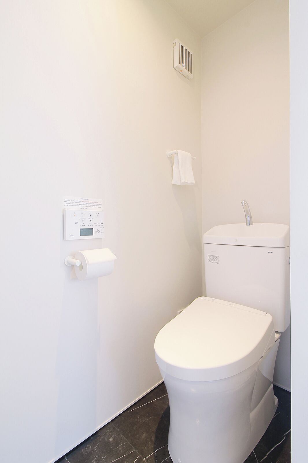 Un baño pequeño y blanco con inodoro, dispensador de toallas de papel y una toalla colgada en la pared.