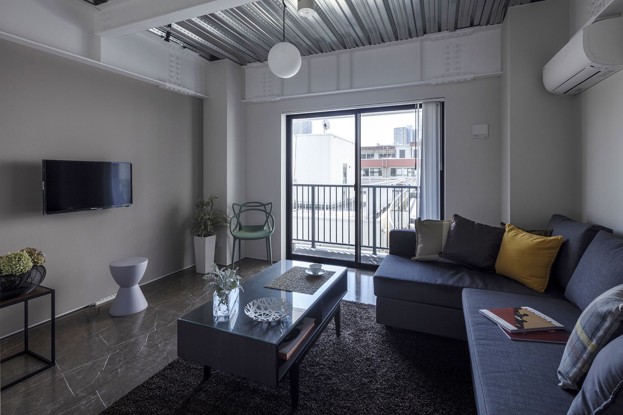 A modern living room with a flat-screen TV, a glass coffee table, a sofa with cushions, a potted plant, and a view of the city through large windows.