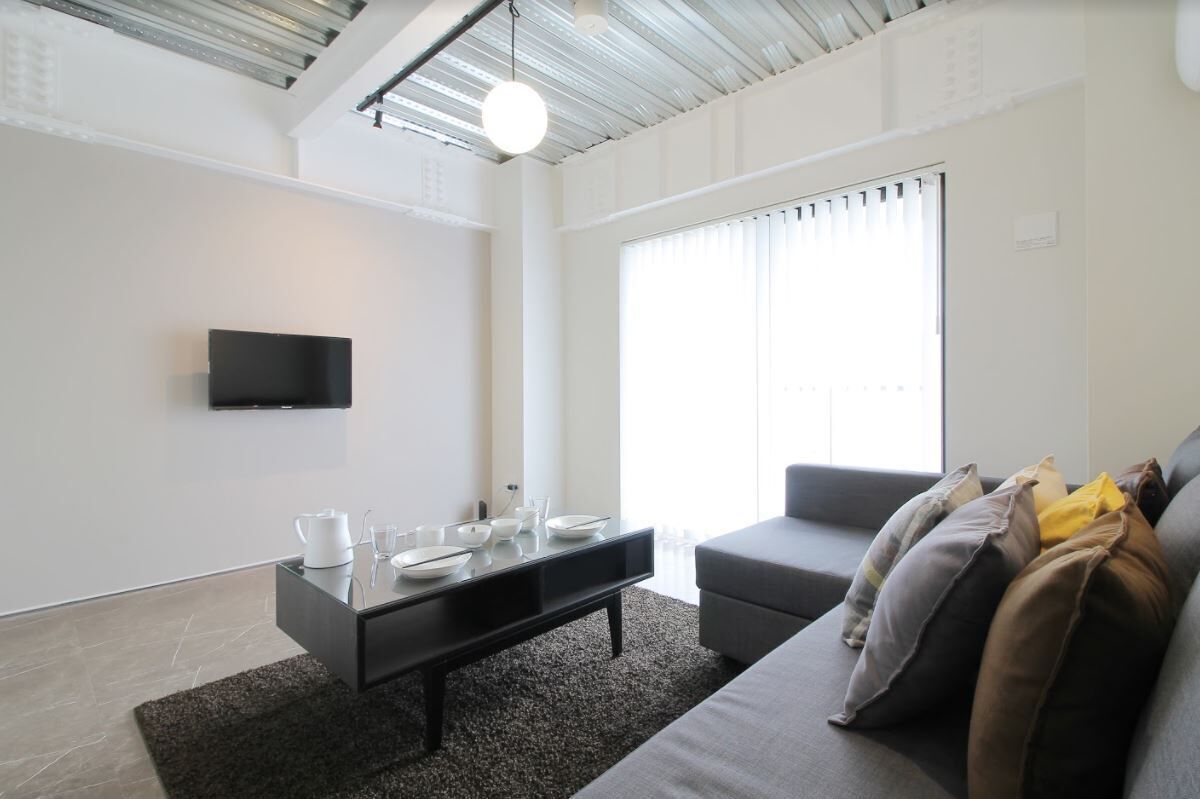 Un salon moderne avec un téléviseur à écran plat fixé au mur, une table basse noire agrémentée de bols et de verres, un canapé gris avec des coussins et une grande fenêtre avec des stores.