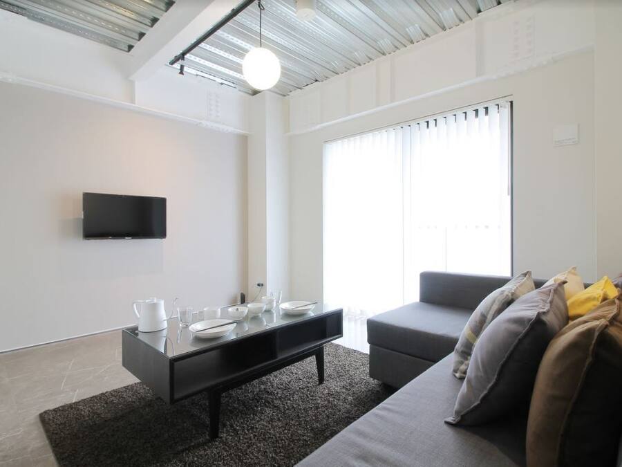 Una sala de estar moderna con un televisor de pantalla plana montado en la pared, una mesa de centro negra con cuencos y vasos, un sofá gris con cojines y un ventanal con persianas.