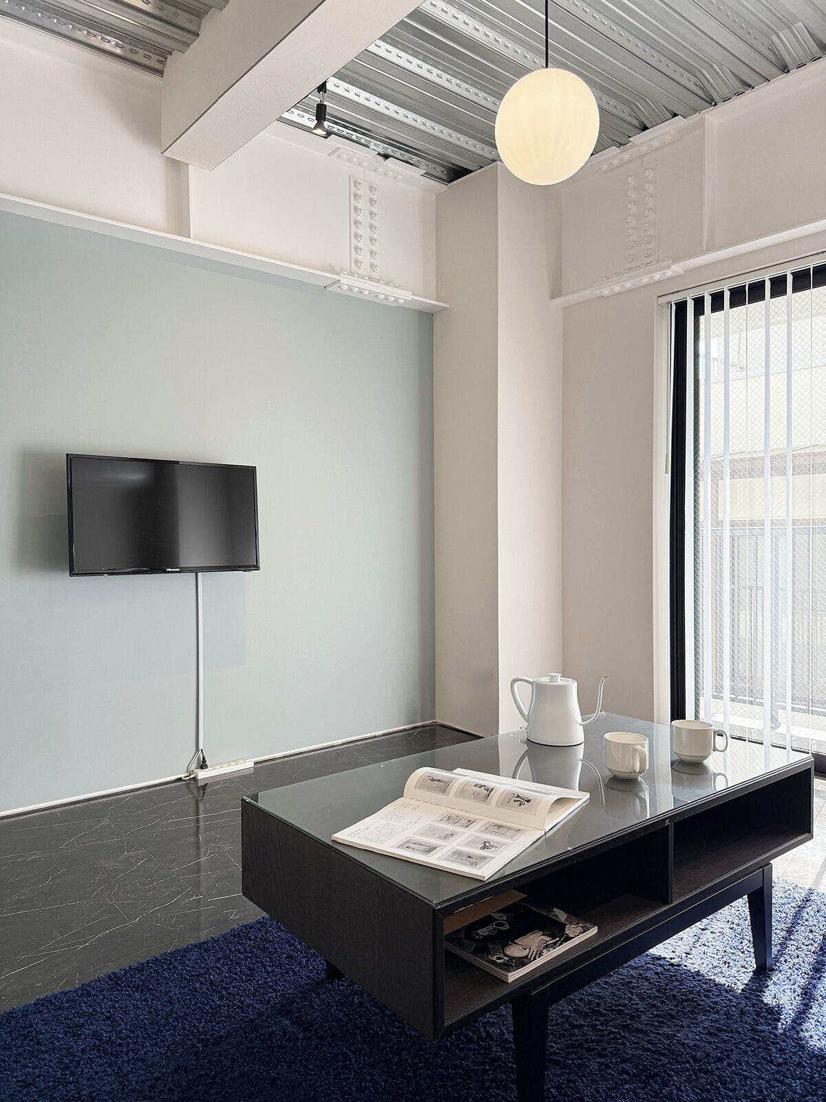 A modern room with a flat-screen TV mounted on the wall, a glass coffee table with a magazine, a kettle, and two cups on a windowsill, and a dark blue carpet.