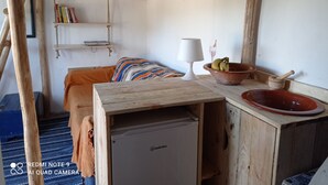 A compact room with a bed, a small table with a lamp, a bowl of fruit, and a mini fridge.