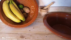 A wooden table with a terracotta bowl containing bananas and potatoes, a wooden mortar and pestle, and a terracotta basin.