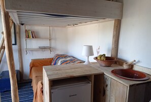 Habitació senzilla amb llit, taula petita amb làmpada, un bol de fruita i nevera petita.