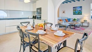 A modern kitchen with white cabinets, a wooden dining table set with chairs, and a view of a living area with a sofa and a painting. - Villa Terrace sur Mer by Interhome (Jávea)