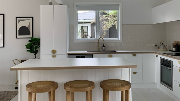 Uma cozinha moderna com armários brancos, uma ilha central com banquetas, uma janela com vista e uma pia com torneira.