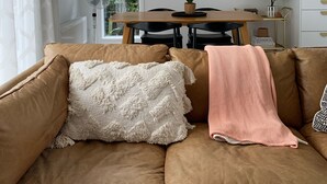 Uma sala de estar com um sofá marrom, uma porta branca, uma mesa de jantar com cadeiras, um pendente de rattan e um armário branco.