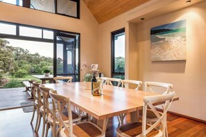 A dining area with a wooden table and chairs, large windows, and a painting of a beach scene.