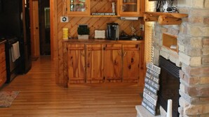 A cabin interior with wooden floors, a stone fireplace, and a kitchen area with wooden cabinets and a ceiling fan.