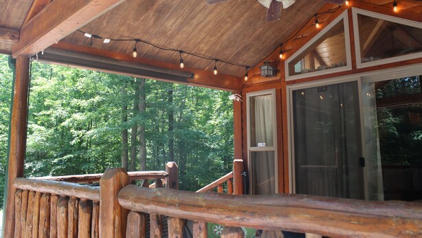 A wooden cabin with a porch, a ceiling fan, string lights, and a view of the forest.