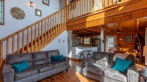 Ein großzügiger Wohnbereich mit zwei grauen Sofas, Holzböden, einer Holztreppe und einer Küche im Hintergrund.