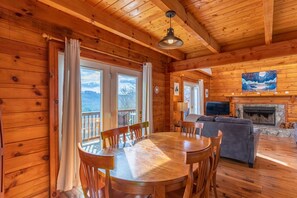 L’intérieur d’un chalet en bois comprend une table à manger, des chaises, un canapé, une cheminée et de grandes fenêtres offrant une vue sur les montagnes.