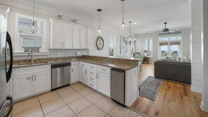 Cocina moderna con armarios blancos, encimeras de granito, electrodomésticos de acero inoxidable, iluminación colgante y vistas al área de estar.