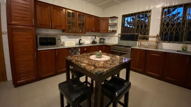 A kitchen with wooden cabinets, a granite countertop, and a central island with four stools. - Cozy 3BR home - gated estate - Tema - top security (Tema)