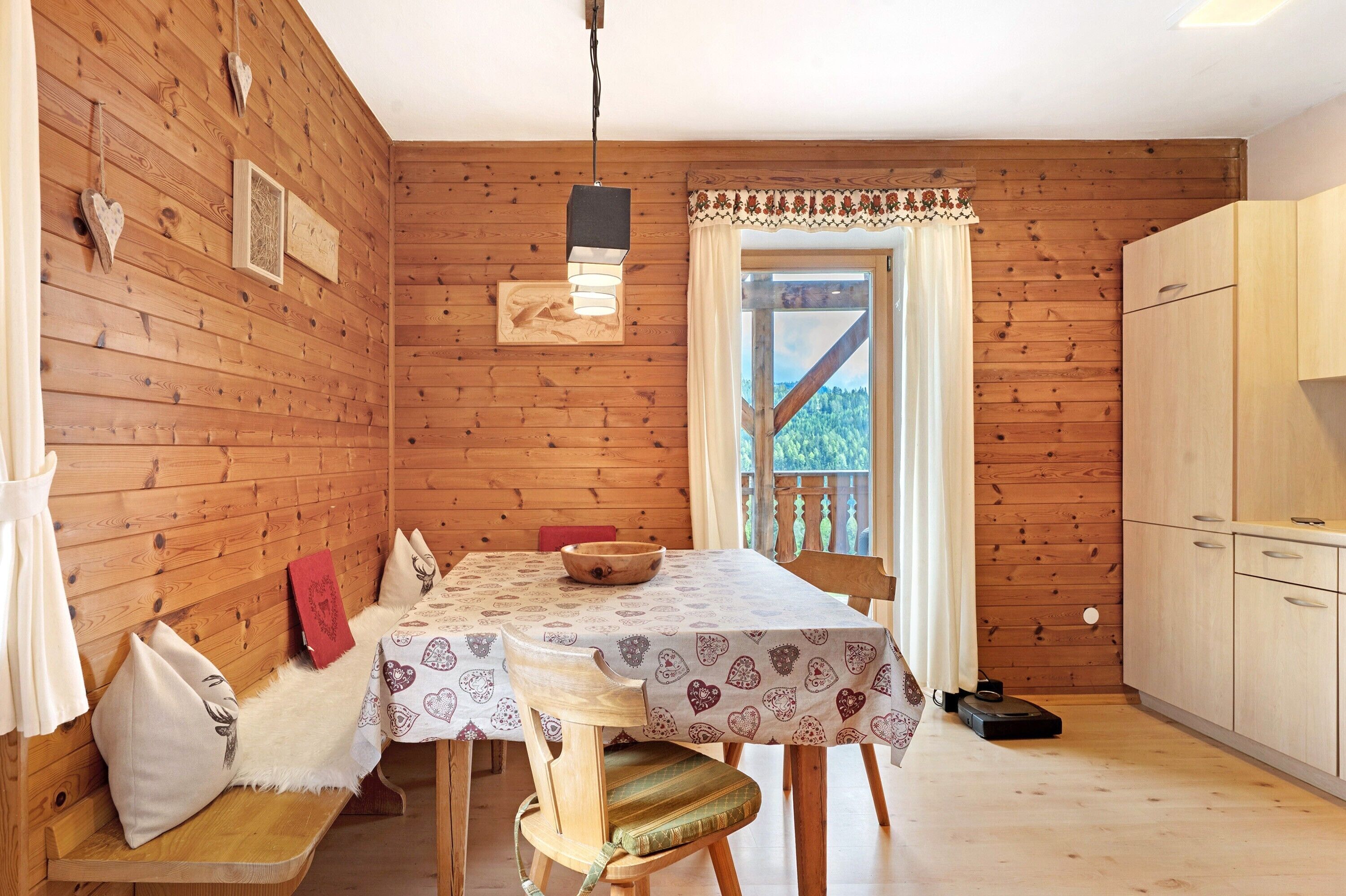 A wooden cabin interior with a dining table, chairs, a bench, a kitchen area, and a window with a view.