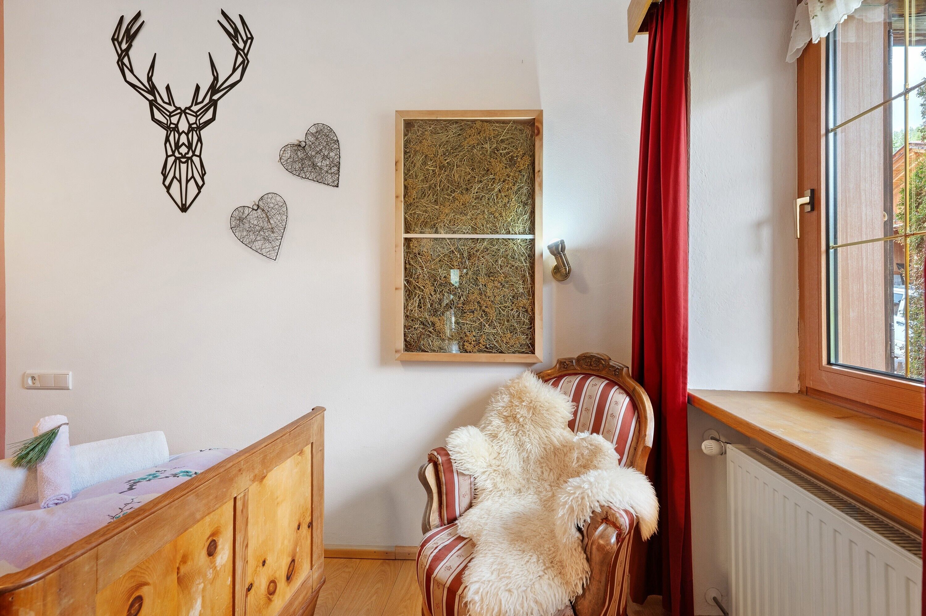 Une pièce comprenant un lit, un fauteuil recouvert d’un plaid en fourrure, une fenêtre donnant sur un paysage agréable, et des éléments de décoration murale, notamment un crâne de cerf et des motifs en forme de cœur.