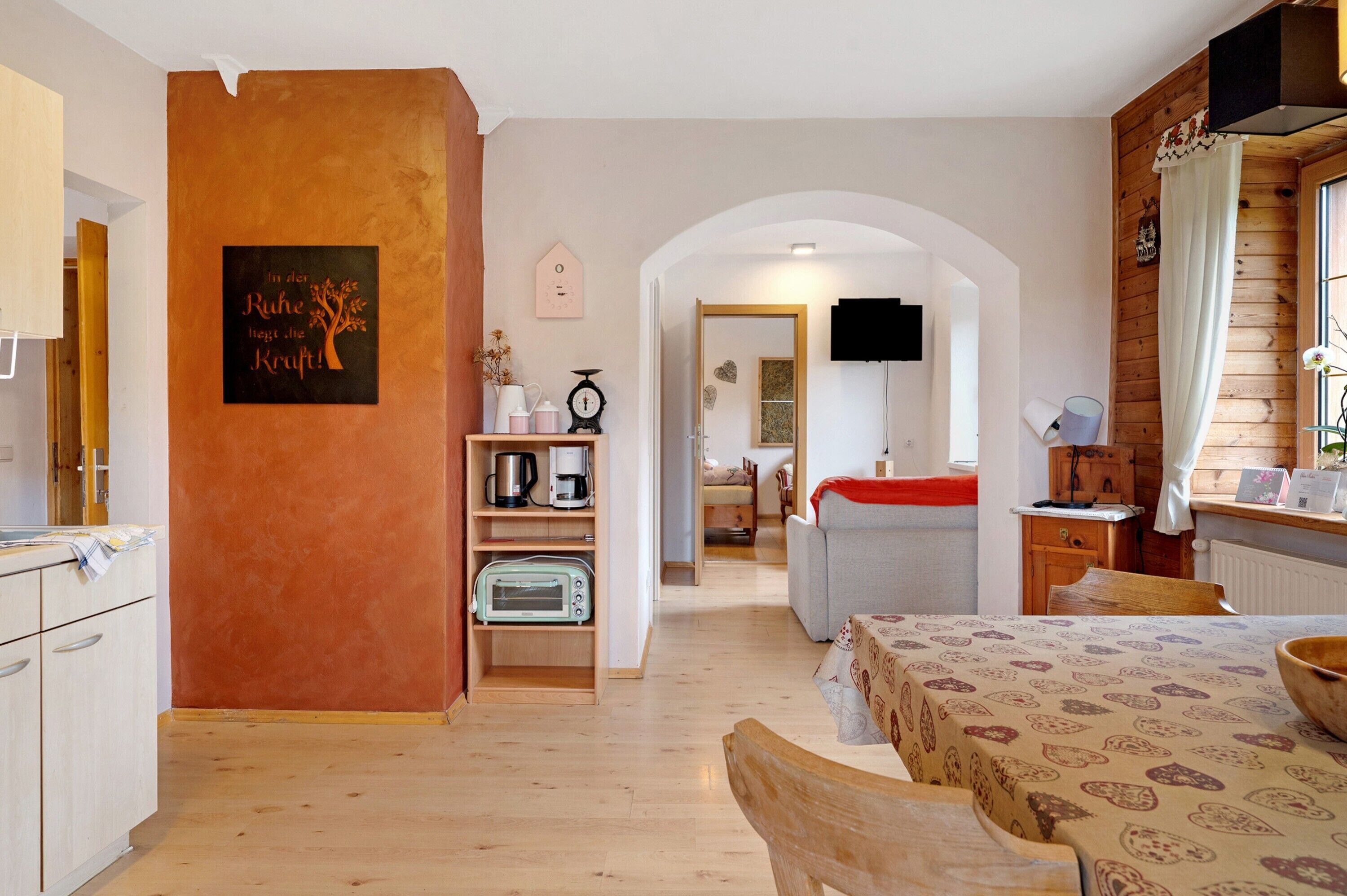 A cozy room with a wooden floor, a dining table with a patterned tablecloth, a kitchen area with a toaster, and a TV mounted on the wall.