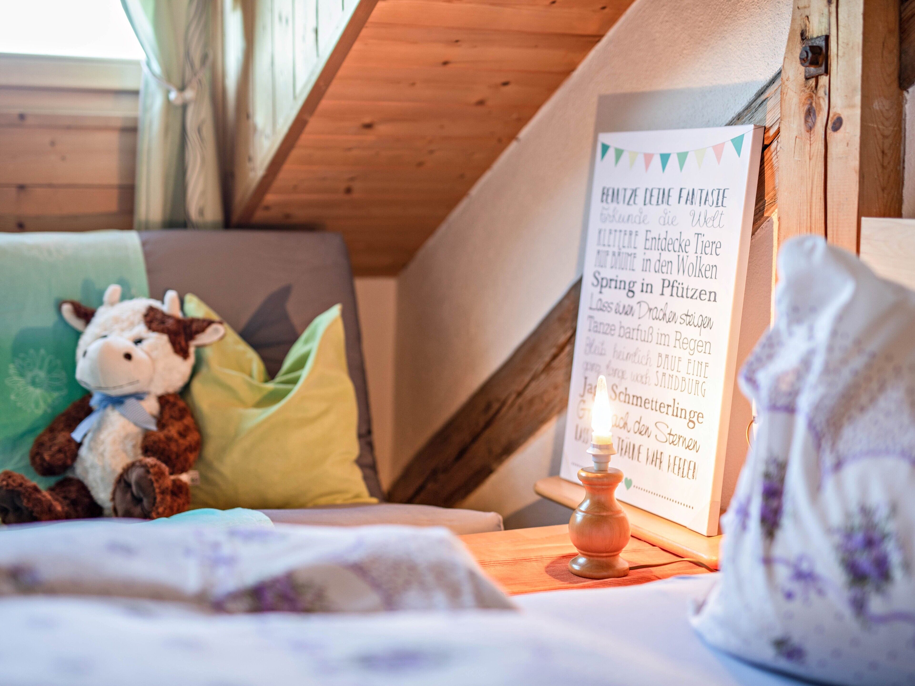 A cozy room with a wooden ceiling, a plush toy, a green pillow, a framed text on the wall, and a small candle on a wooden table.