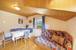 A cozy room with a wooden ceiling, a dining table with chairs, a colorful sofa, and a window with curtains.