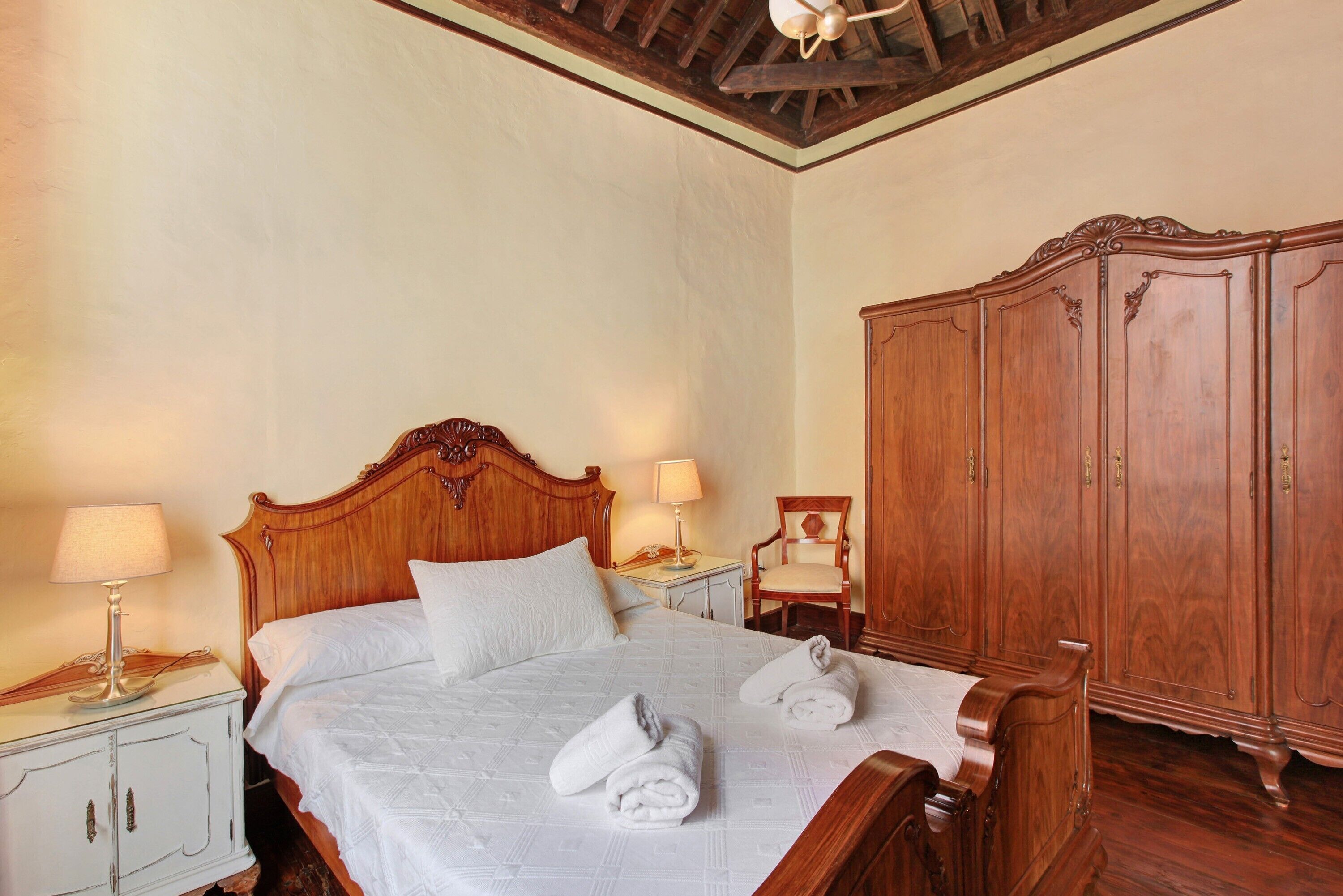 Una cama bien tendida con sábanas y toallas blancas, un armario de madera, una mesita de noche con una lámpara y una silla.
