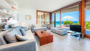 Una sala de estar moderna con un sofá grande, una mesa de centro y un televisor sobre un mueble. Hay un balcón con vista al océano.