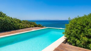 Una piscina rectangular con plataforma de madera, rodeada de vegetación exuberante y vistas despejadas al mar y montañas distantes.