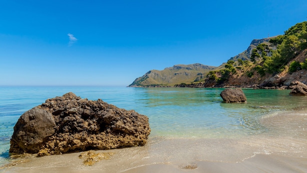 Una playa rocosa con agua cristalina y una montaña al fondo.