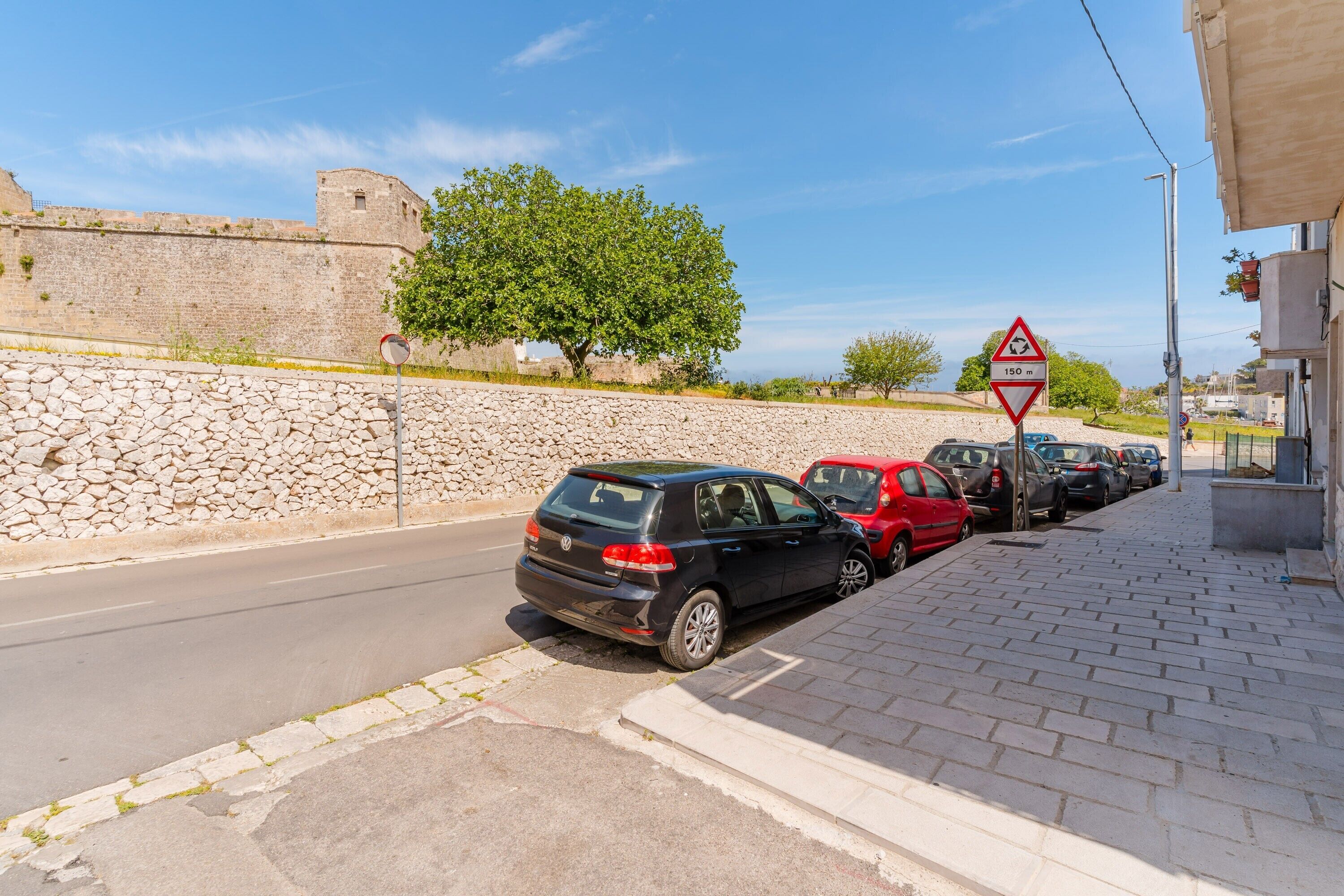 Une rue bordée de voitures garées, un mur de pierre et un panneau indiquant une dénivellation de 150 mètres.