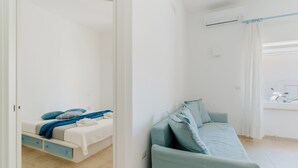 A bedroom with a bed, a blue sofa, a window with curtains, and a door leading to another room.