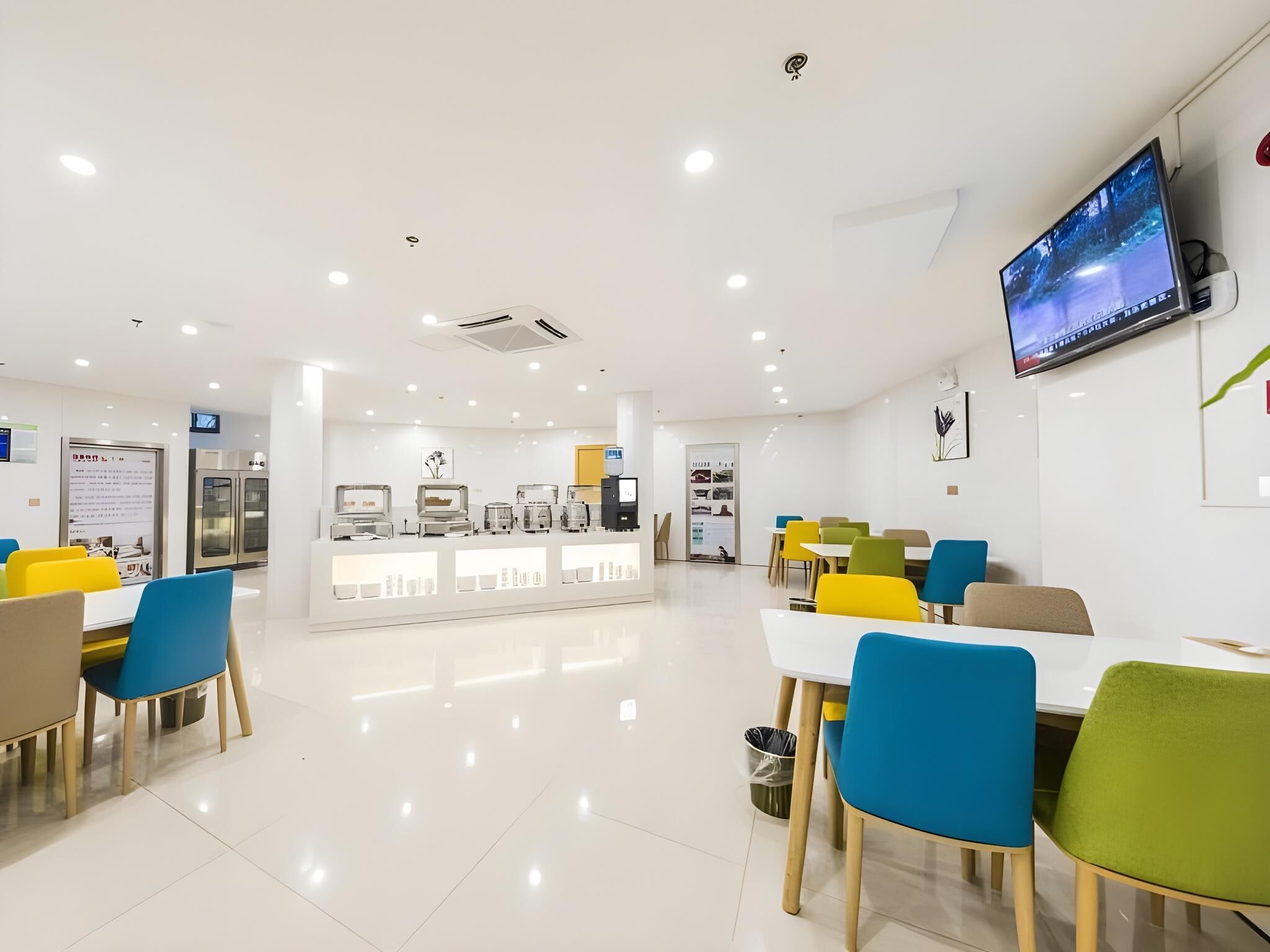 Un restaurante moderno y bien iluminado, con un diseño limpio y minimalista. Cuenta con una barra central que exhibe diversos alimentos, rodeada de mesas y sillas de diferentes colores. Un televisor está montado en la pared, y hay cuadros enmarcados adornando las paredes.