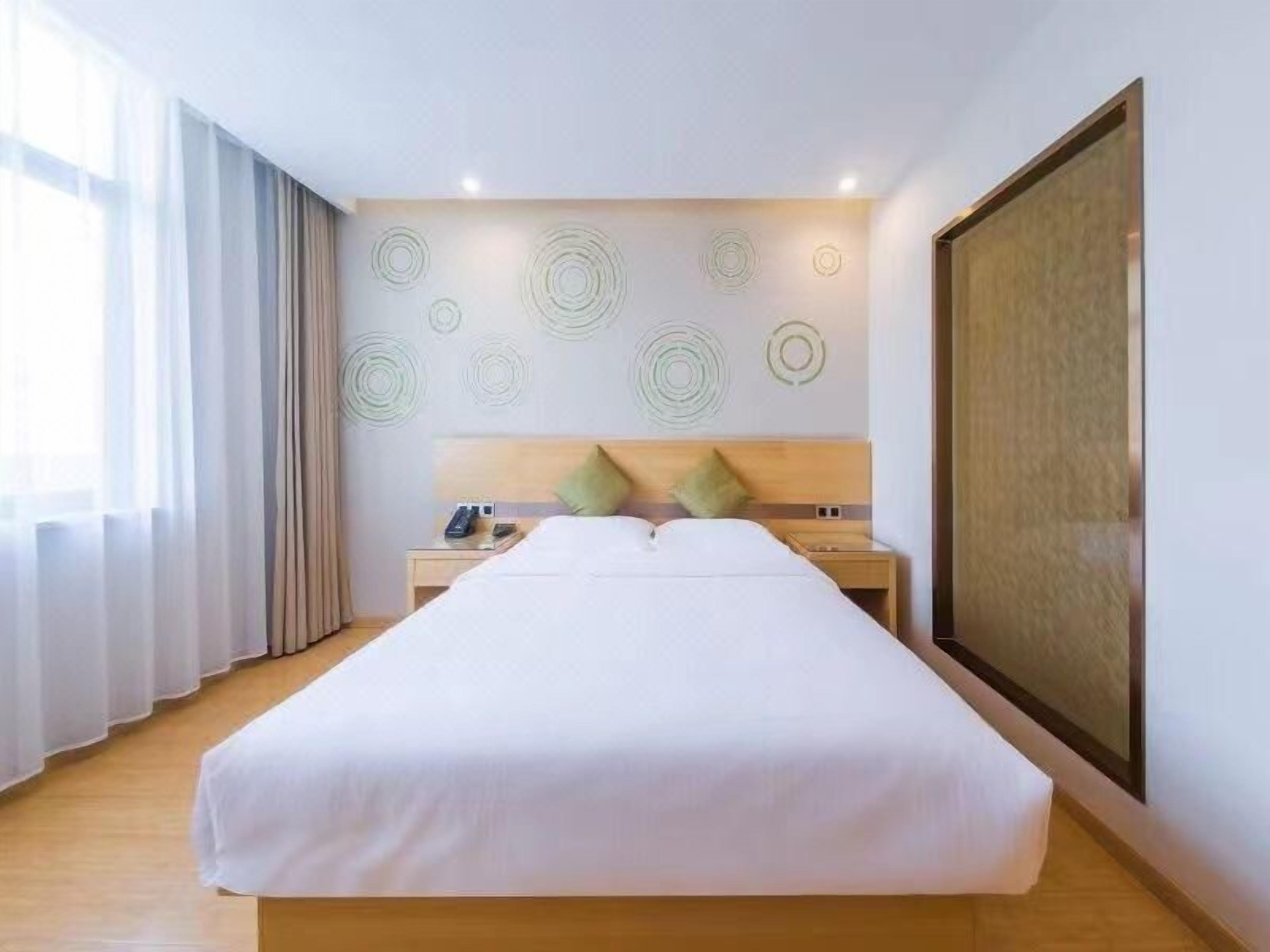 A hotel room with a large bed, a wooden headboard, a nightstand with a phone and remote, and a large window with curtains.