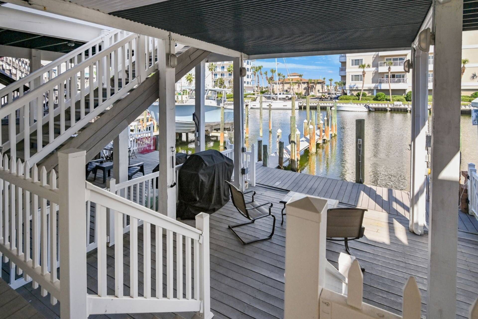 Una terraza techada con vista a un puerto deportivo, que incluye una escalera de madera, mobiliario de exterior y una parrilla.