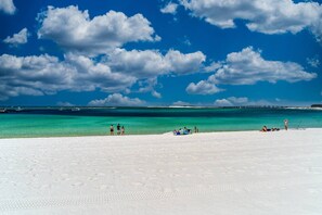 Uma praia com areia branca, água turquesa límpida e céu azul com nuvens.