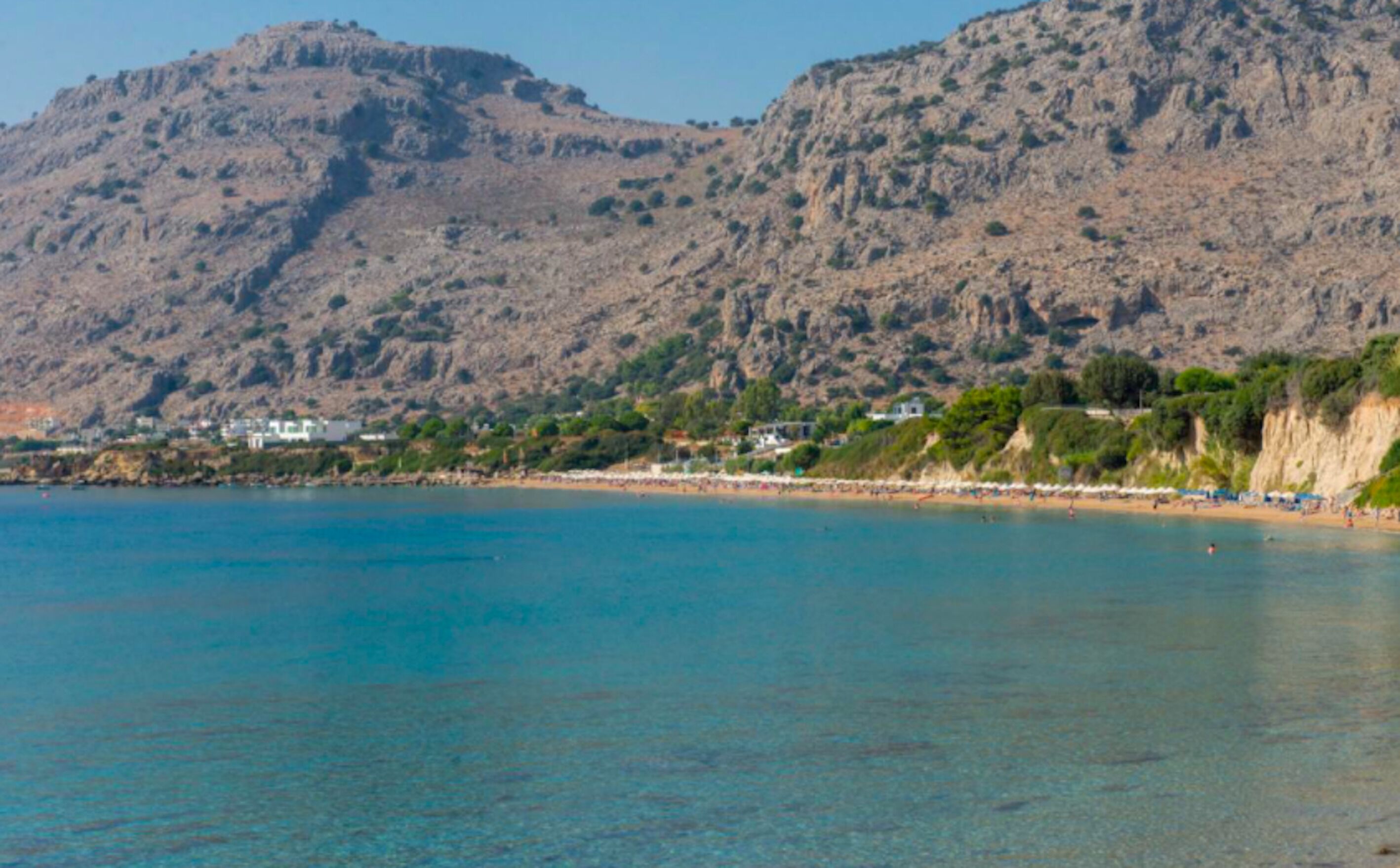 A coastal landscape with a clear blue sea, rocky cliffs, and a beach with buildings along the shore.