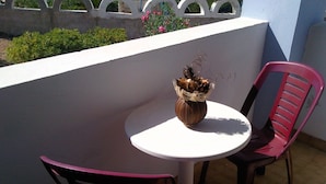 Una pequeña mesa al aire libre con un florero de flores secas, dos sillas de plástico y una pared blanca con una cerca decorativa al fondo.