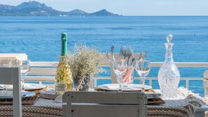 A table set with dinnerware, a bottle of champagne, and a crystal decanter, overlooking a coastal view with mountains in the distance.