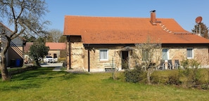 A two-story stone house with a red-tiled roof, a visible chimney, and a small front porch with outdoor seating.