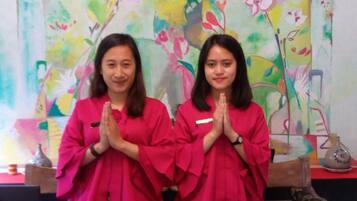 Two individuals in red robes seated at a wooden table with a colorful floral painting in the background.
