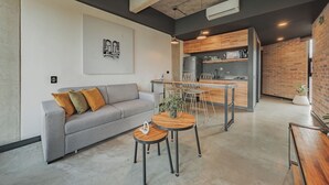 Een moderne woonruimte met een grijze bank, twee kleine ronde tafels en een keuken met houten kastjes en een roestvrijstalen koelkast.