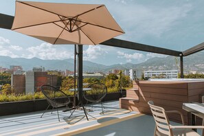 Een terras op het dak met een grote parasol, twee stoelen, een tafel en een houten bankje.