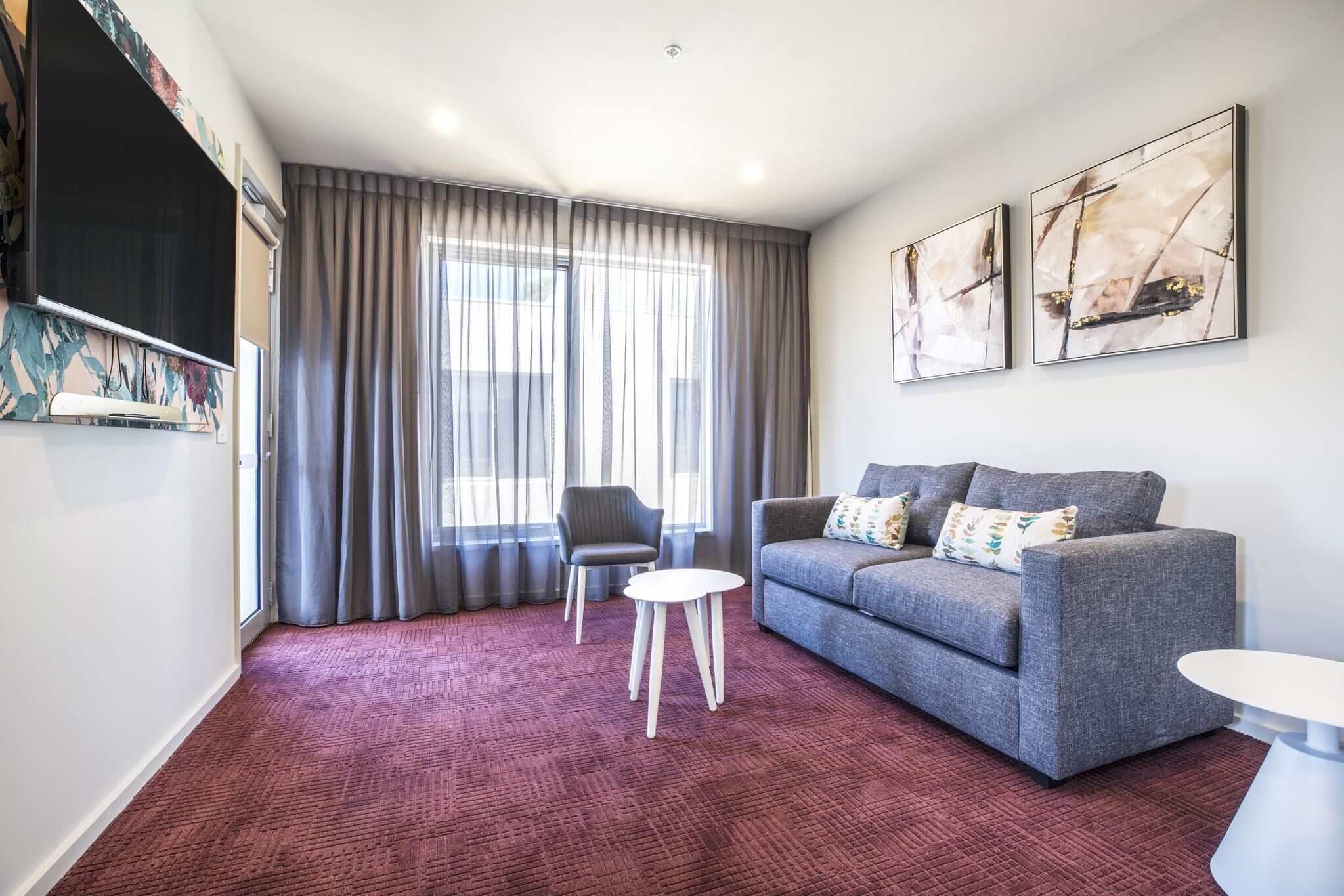 Ein modernes Wohnzimmer mit einem grauen Sofa, einem kleinen weißen Tisch und einem grauen Sessel. An der Wand hängen zwei abstrakte Gemälde und ein großes Fenster mit Vorhängen.